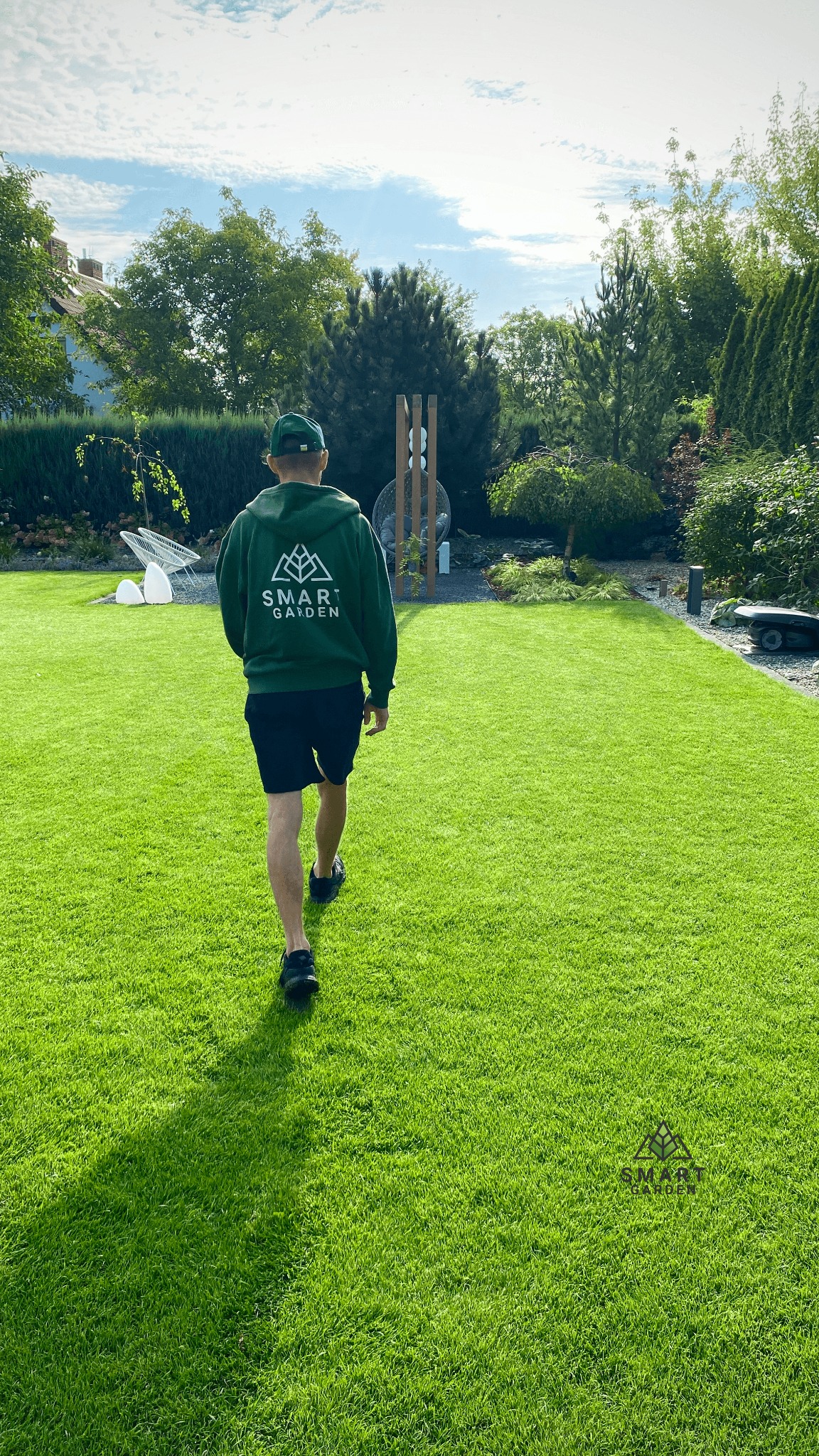 Mężczyzna w zielonej bluzie z logo firmy spaceruje po idealnie przystrzyżonym, zielonym trawniku w ogrodzie. W tle widoczne drzewa i elementy wyposażenia ogrodu, w tym robot koszący.