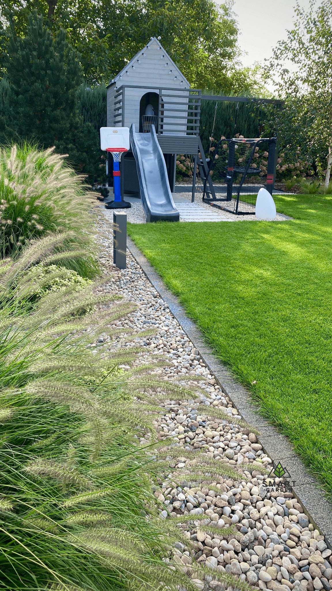 Nowoczesny ogród z domkiem na drzewie, zjeżdżalnią i boiskiem do koszykówki, obrzeża z kamieni i ozdobne trawy, zielony trawnik.