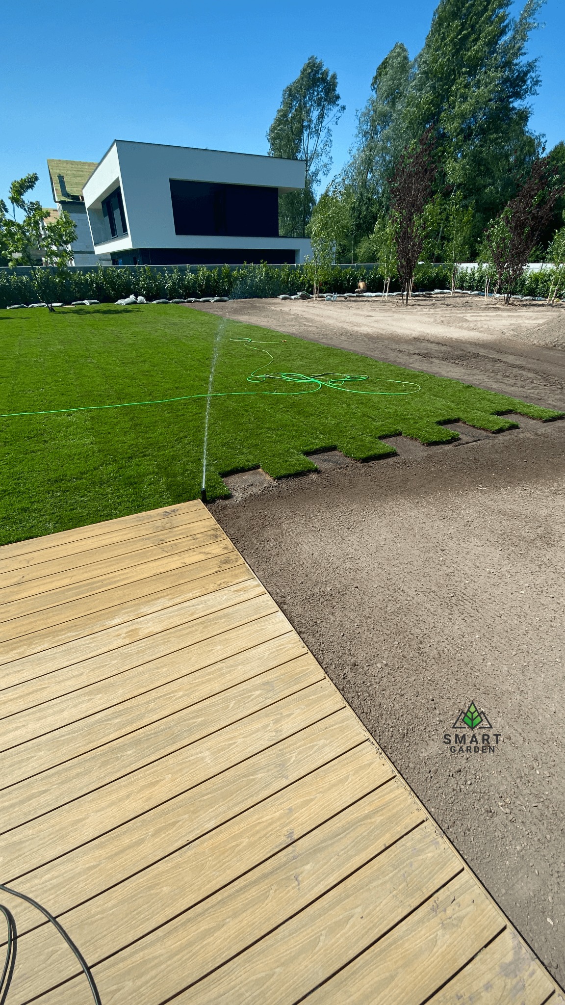 Układanie trawy z rolki w ogrodzie nowoczesnego domu, widok z drewnianego tarasu, system nawadniania w trakcie pracy.