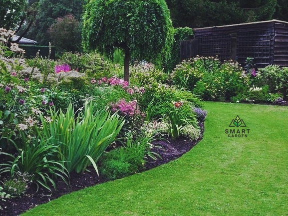 Bujny, kwitnący ogród z drzewem o kulistej koronie i różnorodnymi kwiatami, otoczony równo przystrzyżonym trawnikiem, z drewnianą konstrukcją w tle.