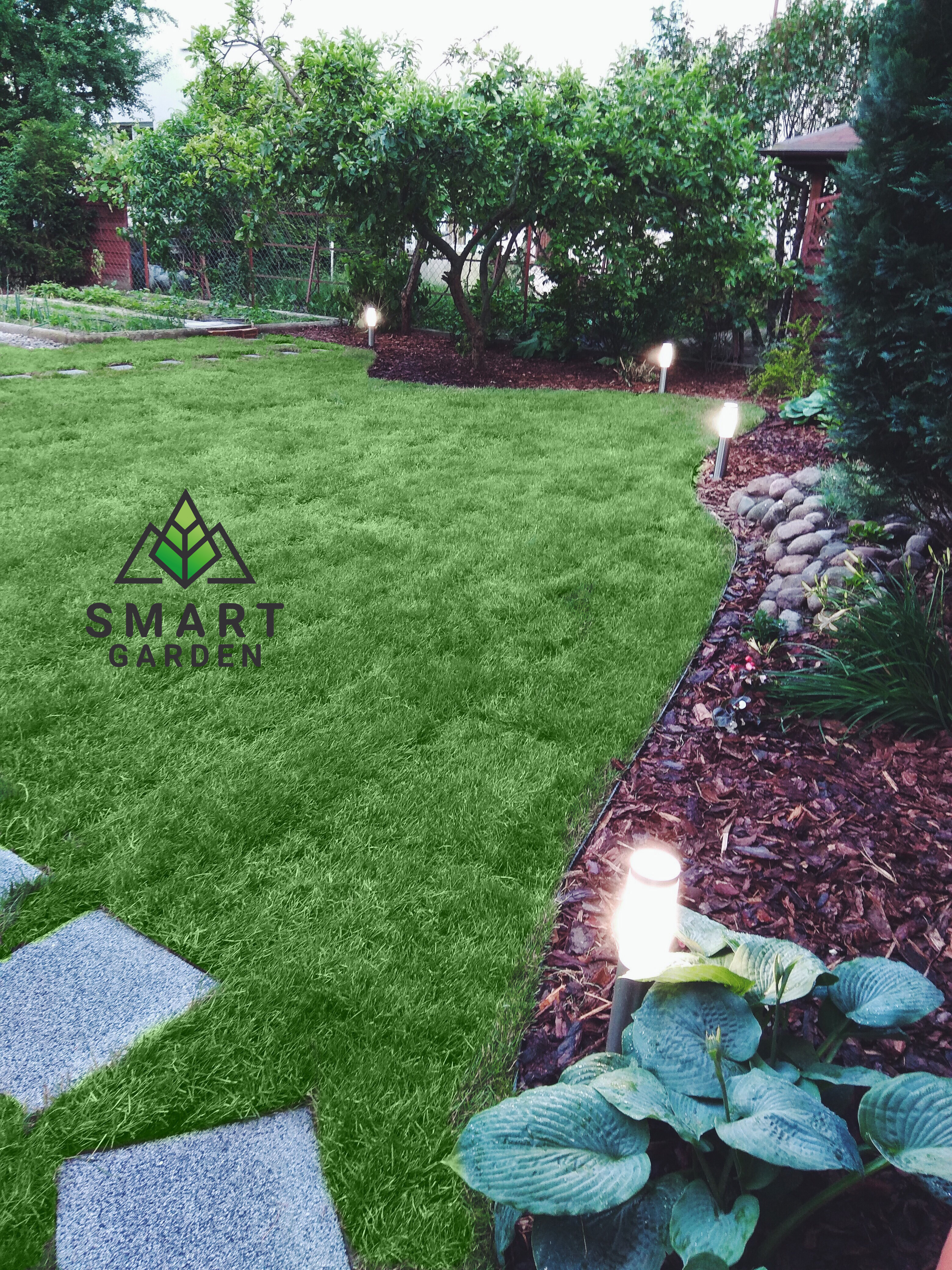 Ujęcie ogrodu z równo przystrzyżonym trawnikiem, ścieżką z płyt, rabatami z korą i kamieniami oraz oświetleniem ogrodowym w postaci niskich lamp, z logo firmy na trawniku.