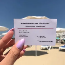 Wizytówka biura rachunkowego 'Rozliczeni' trzymana w dłoni z pomalowanymi paznokciami na tle plaży z parasolami i leżakami.