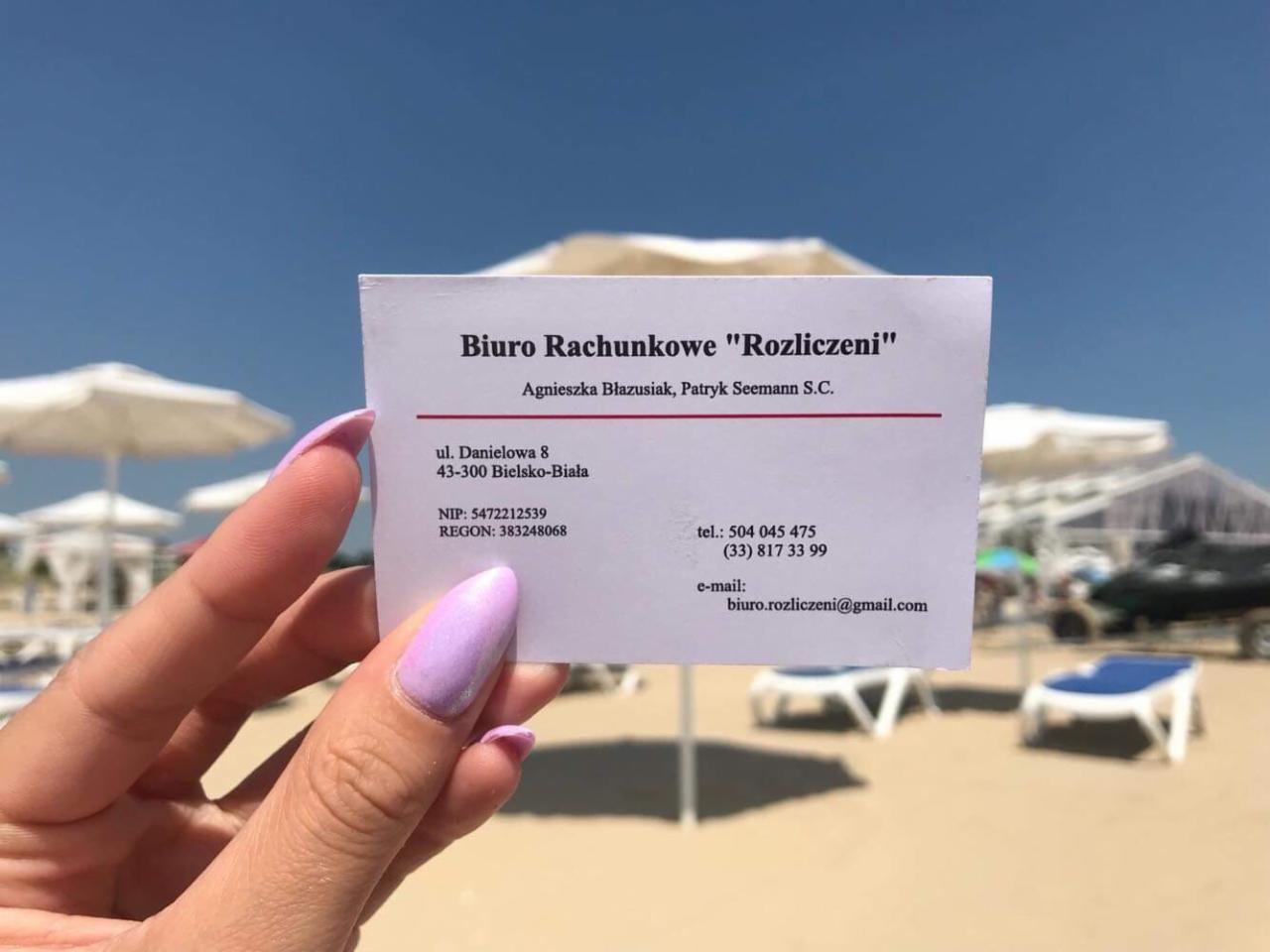 Wizytówka biura rachunkowego 'Rozliczeni' trzymana w dłoni z pomalowanymi paznokciami na tle plaży z parasolami i leżakami.