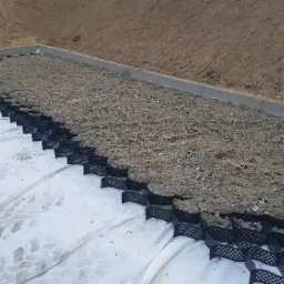 Geowłóknina z czarną geokratą i wypełnieniem gruntem, obrzeże z kostki betonowej na skraju wykopu.