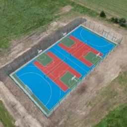 Widok z lotu ptaka na nowo wybudowane, wielofunkcyjne boisko sportowe z nawierzchnią w kolorach niebieskim, czerwonym i zielonym, otoczone siatką i świeżo wyrównanym terenem.