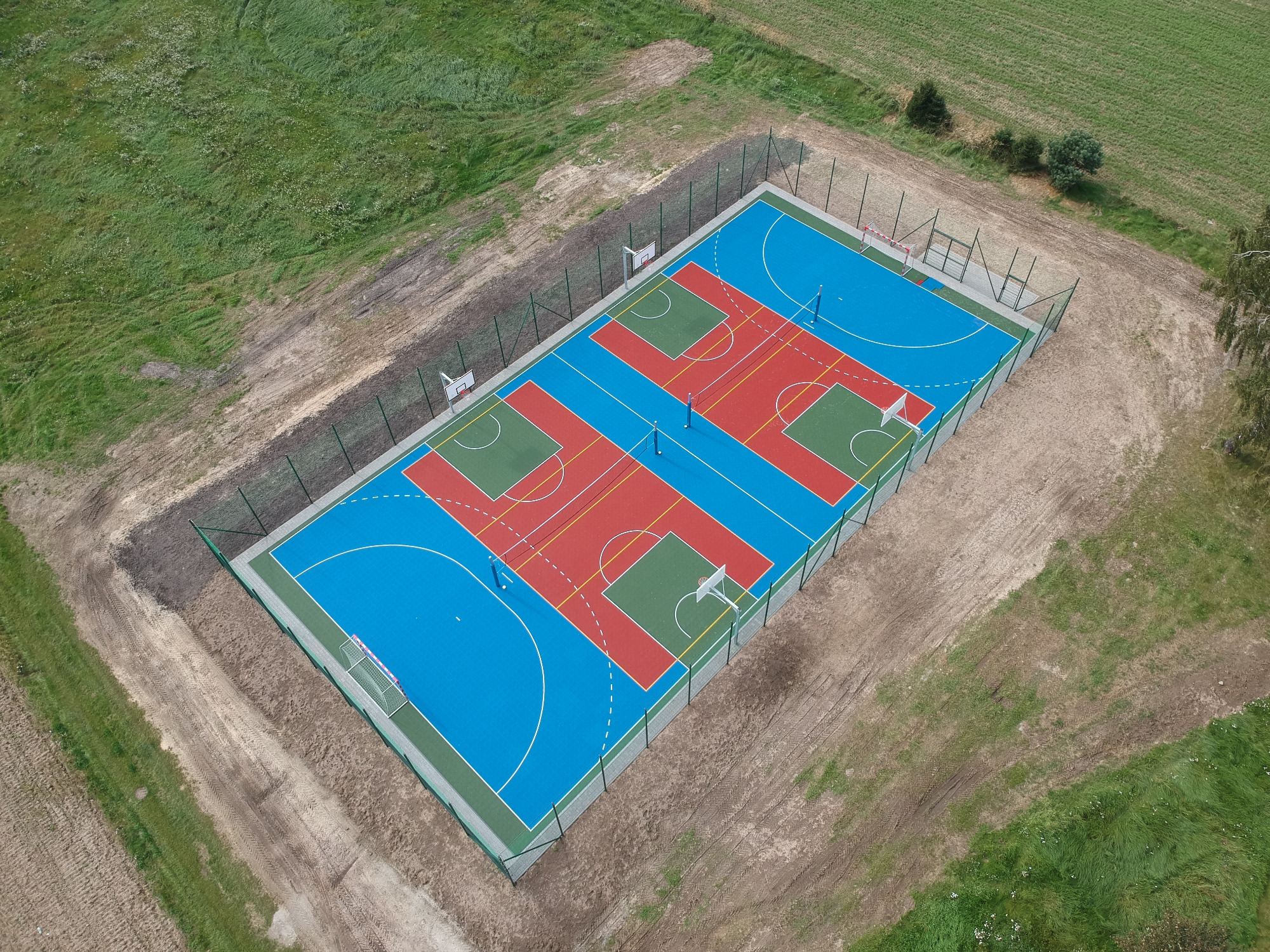 Widok z lotu ptaka na nowo wybudowane, wielofunkcyjne boisko sportowe z nawierzchnią w kolorach niebieskim, czerwonym i zielonym, otoczone siatką i świeżo wyrównanym terenem.