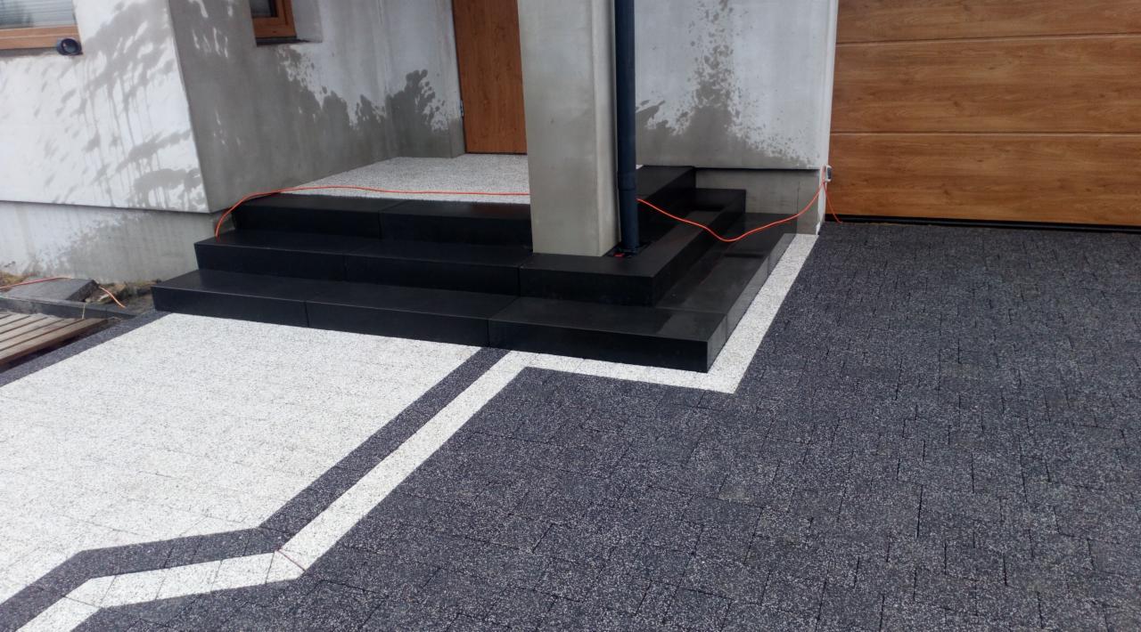 Nowoczesne schody wejściowe z czarnego granitu, kontrastujące z jasną kostką na podjeździe, tworząc geometryczny wzór z ciemnoszarej kostki. Widoczny fragment elewacji budynku z drewnianą bramą...