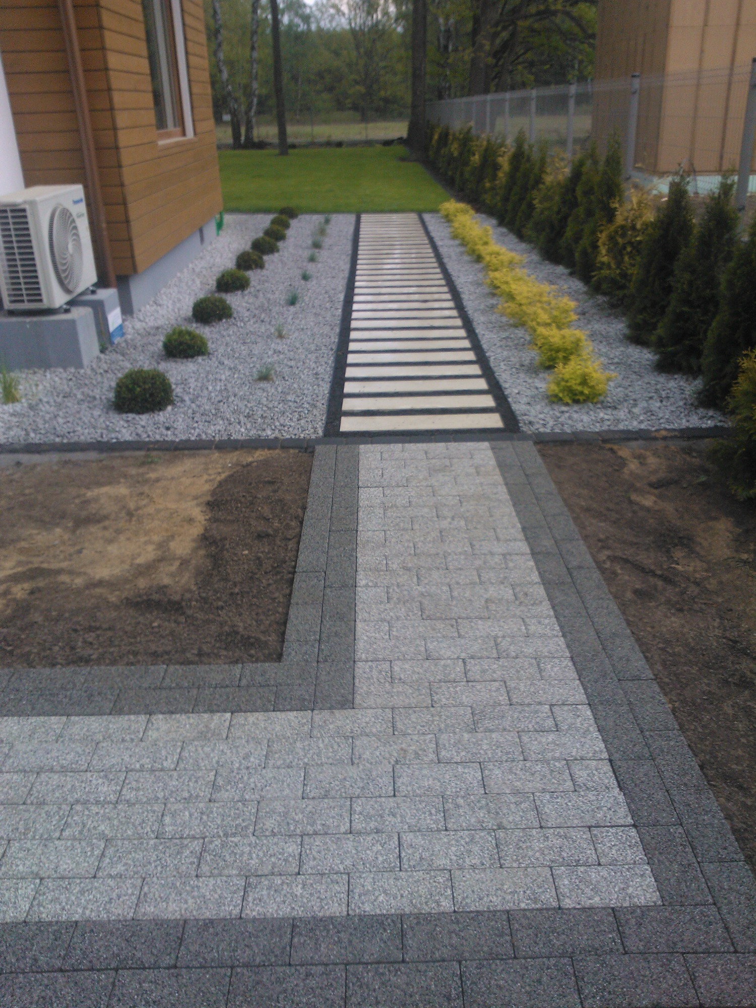 Ścieżka z jasnoszarej kostki granitowej z ciemnoszarą obwódką, przechodząca w ścieżkę z płyt betonowych, otoczona ozdobnym żwirem i roślinnością, widok z góry.