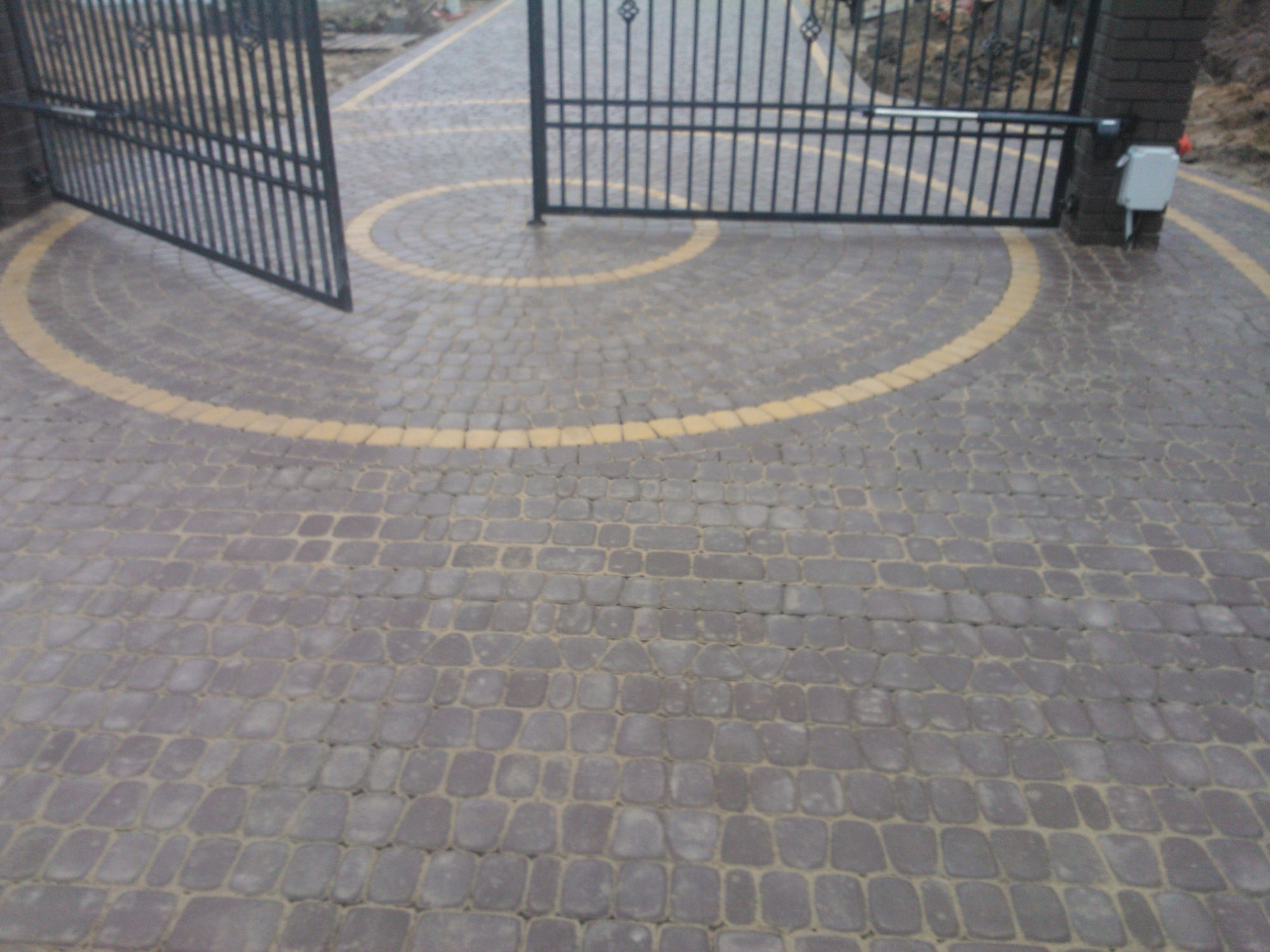 Ułożona kostka brukowa w odcieniach szarości z żółtymi akcentami, tworząca okrąg wokół bramy wjazdowej z automatycznym systemem otwierania.
