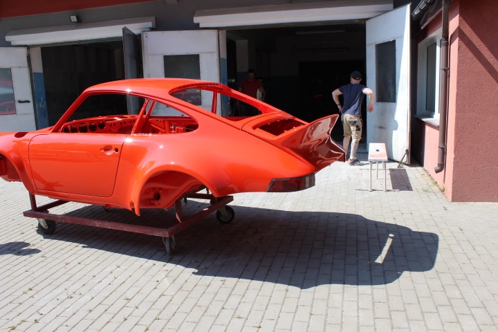Pomarańczowe nadwozie klasycznego Porsche 911 na wózku warsztatowym przed otwartym warsztatem, widoczni mechanicy w tle.