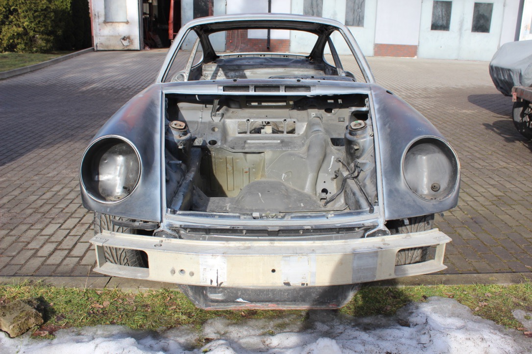 Przód rozebranego samochodu sportowego, przygotowanego do prac renowacyjnych karoserii, z widocznymi elementami nadwozia w szarym podkładzie i fragmentami ośnieżonej trawy u podstawy.