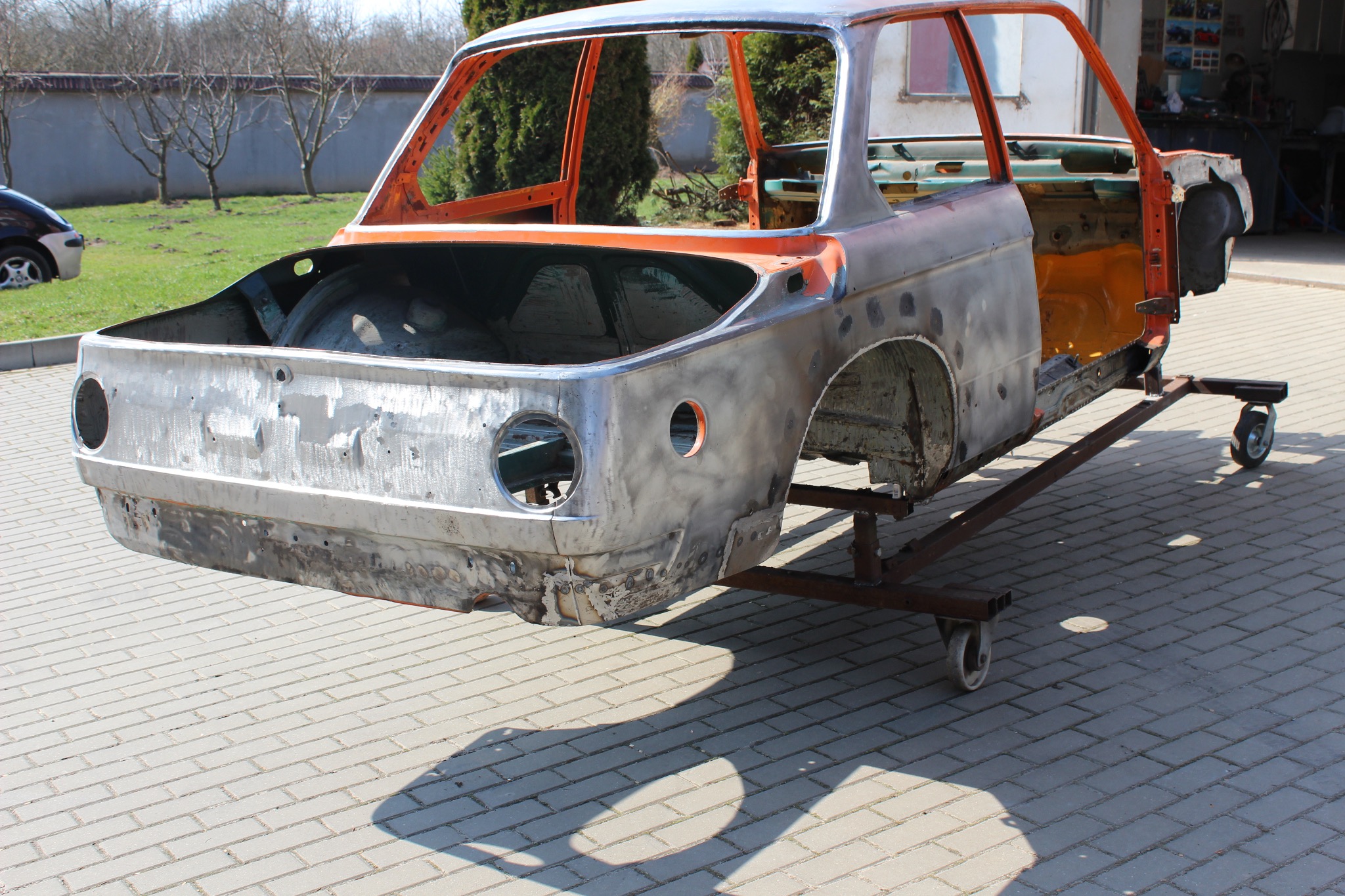 Karoseria samochodu po blacharce, przygotowana do lakierowania, osadzona na mobilnym stojaku na kostce brukowej, z widocznymi śladami spawania i szlifowania.