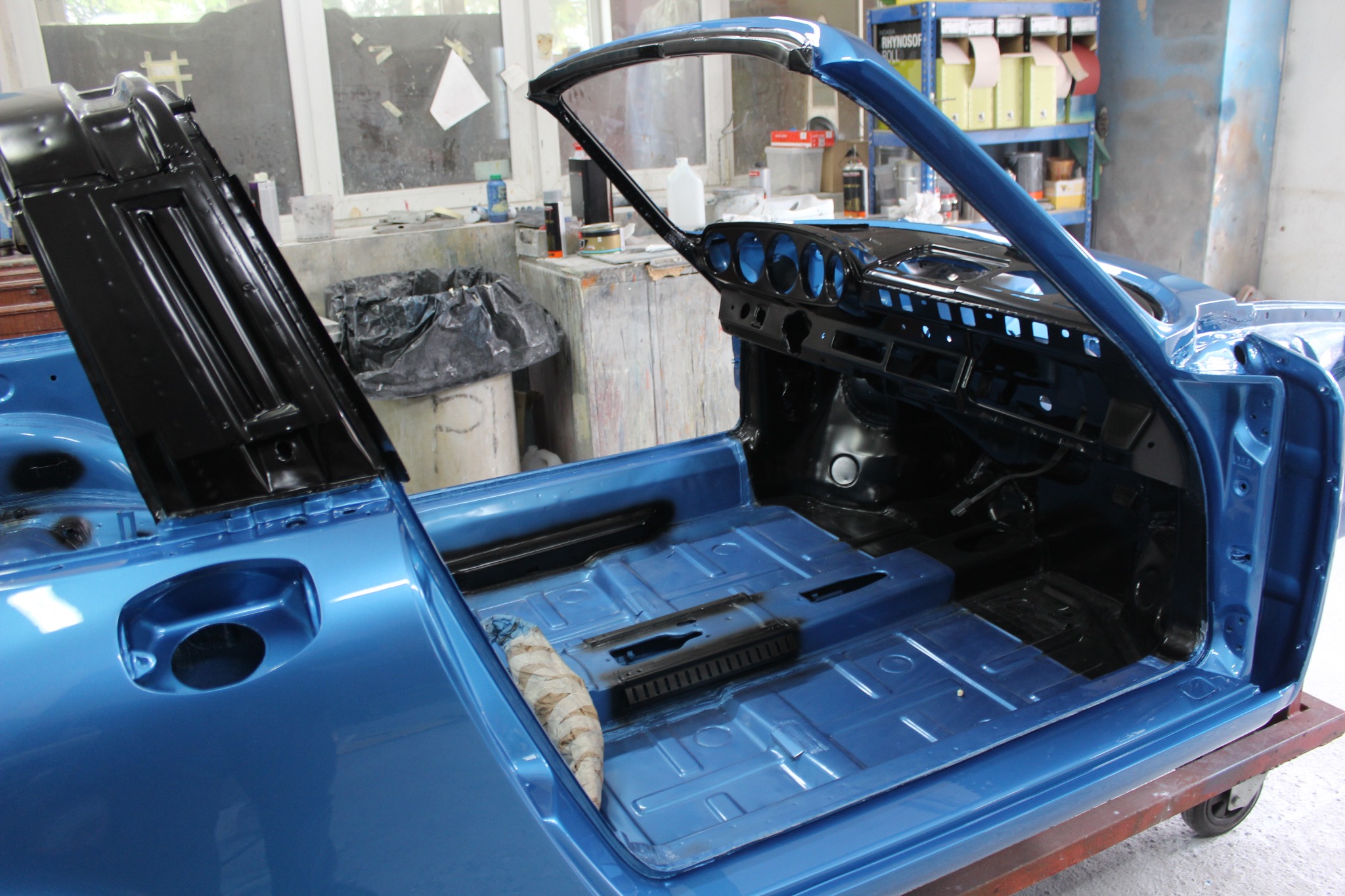Błękitny kabriolet w trakcie renowacji, widoczne wnętrze bez tapicerki i elementy karoserii po lakierowaniu, na warsztatowym wózku.