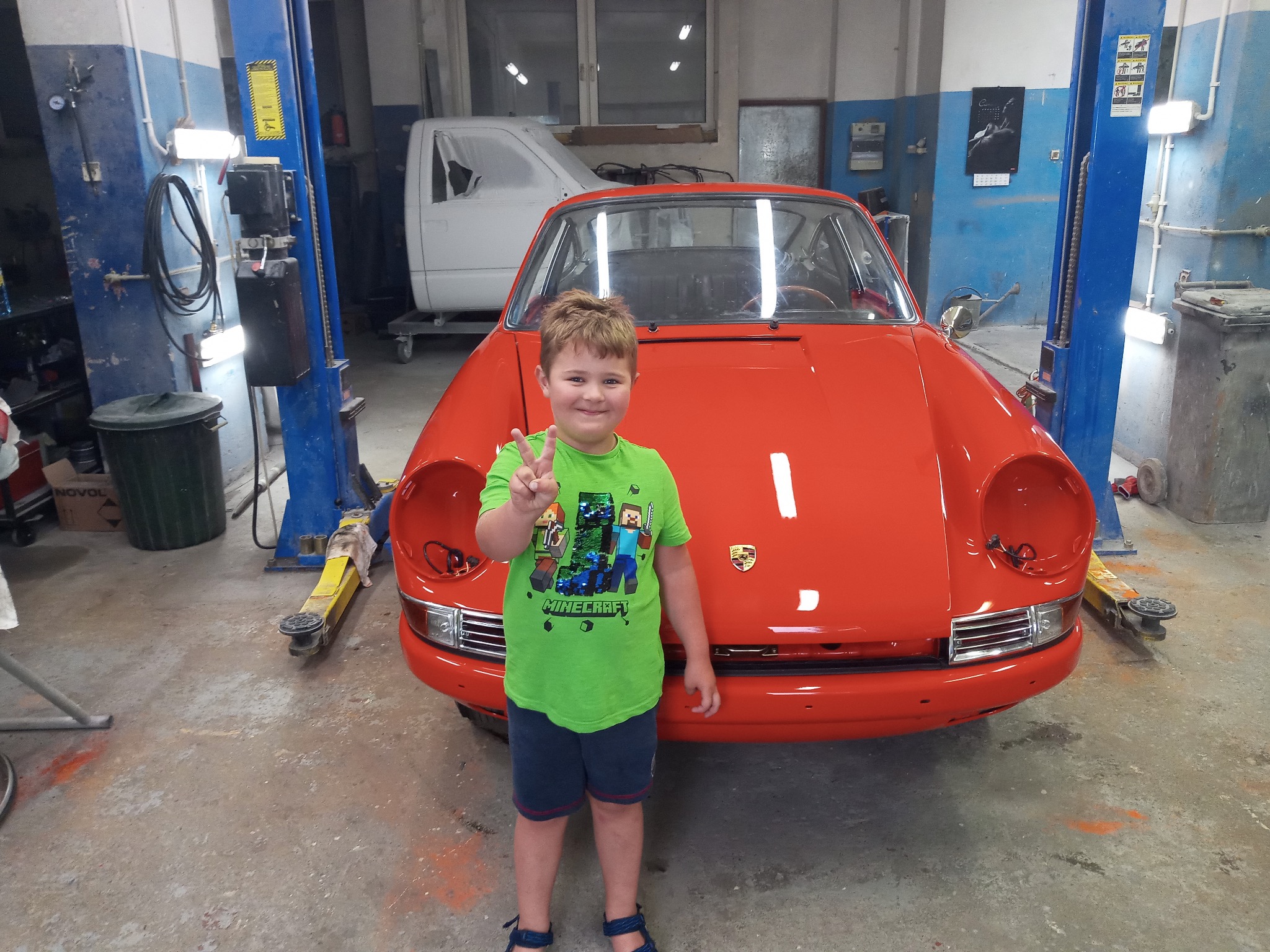 Chłopiec w koszulce Minecraft stoi przed odrestaurowanym, pomarańczowym Porsche 911 na podnośniku w warsztacie.