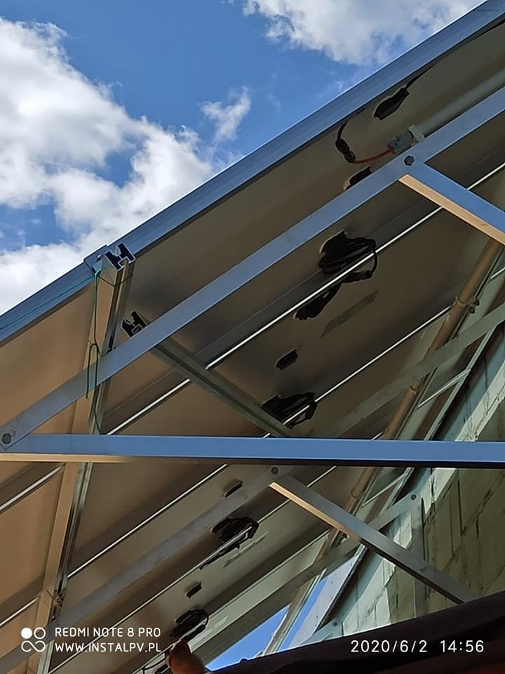 Montaż paneli słonecznych widziany od spodu, z widocznymi elementami konstrukcyjnymi, okablowaniem i fragmentem dachu na tle błękitnego nieba z chmurami.