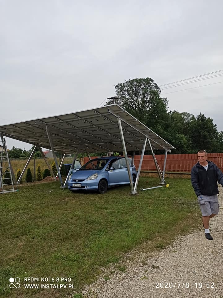 Instalacja fotowoltaiczna jako wiata garażowa nad niebieskim samochodem, widoczny instalator i drabina, trawnik i ogrodzenie w tle.