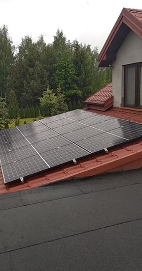 Panele słoneczne zamontowane na dwuspadowym dachu z czerwonej dachówki, widok z góry, w tle drzewa