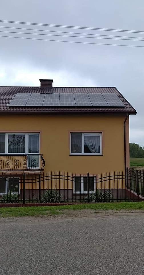 Żółty dom jednorodzinny z ciemnymi panelami słonecznymi na brązowym dachu, widok z ulicy przez czarne metalowe ogrodzenie.