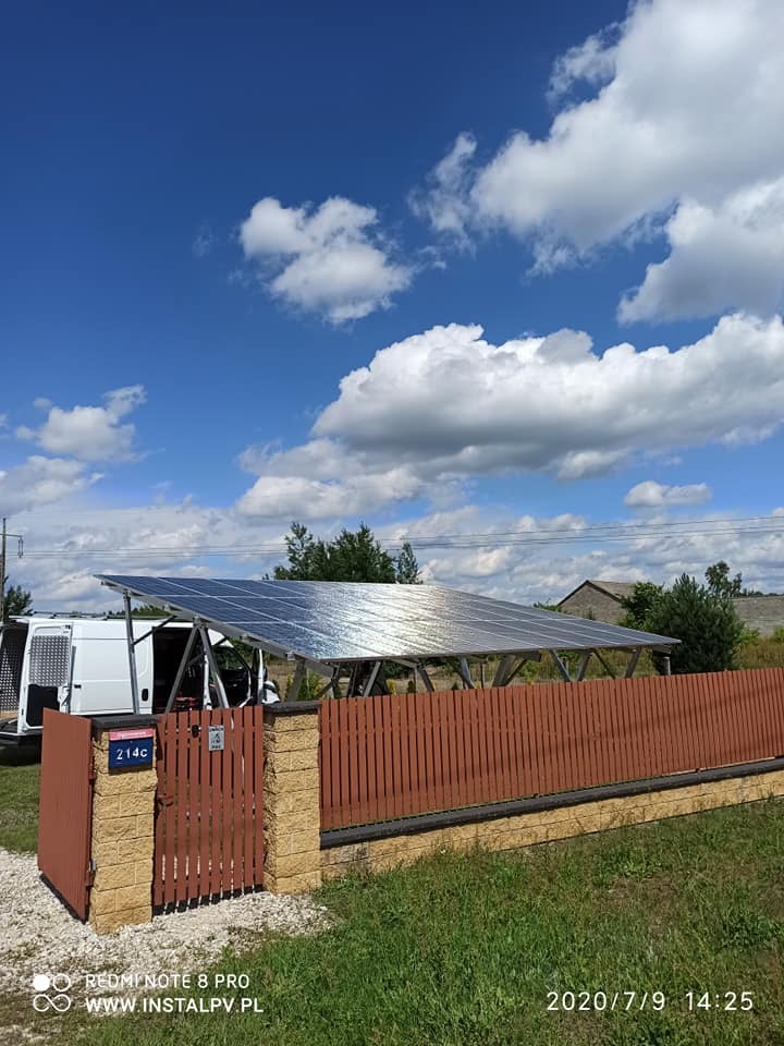 Instalacja paneli słonecznych nad miejscem parkingowym, widoczna zza drewnianego ogrodzenia z numerem domu 214c, z białym vanem w tle i błękitnym niebem z chmurami.
