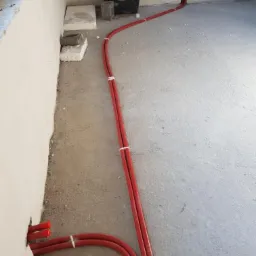 Czerwone rury instalacyjne, spięte białymi opaskami, prowadzone po surowym betonowym podłożu wzdłuż ściany w trakcie montażu.