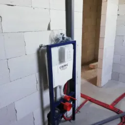 Stelaż podtynkowy WC marki Grohe zamontowany przy ścianie z białego betonu komórkowego, widoczne rury doprowadzające wodę i odpływ kanalizacyjny w surowym stanie.