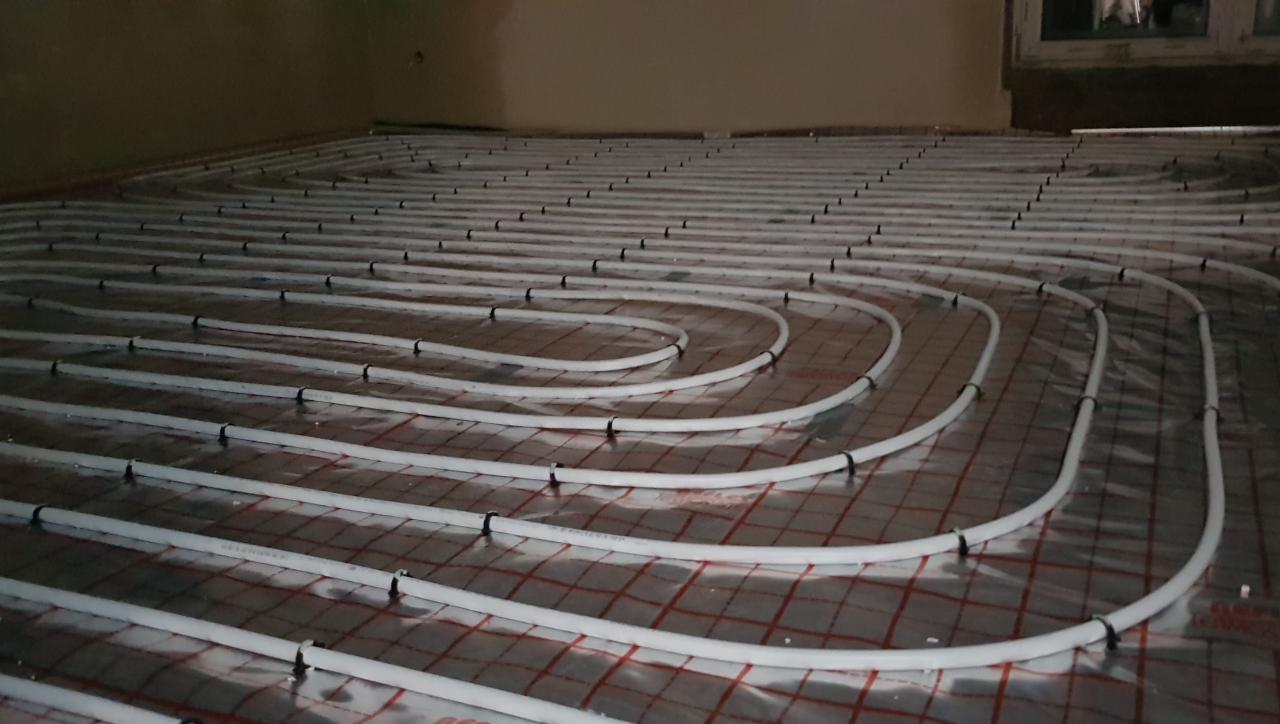 Białe rury systemu ogrzewania podłogowego zamocowane do folii z czerwoną siatką na podłożu, widoczne czarne opaski mocujące.