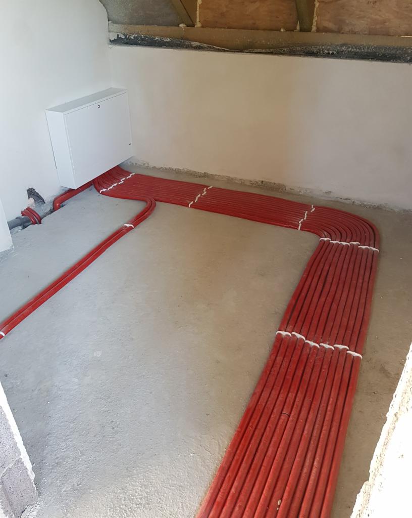 Czerwone rury ułożone równolegle na betonowej podłodze w surowym wnętrzu, prowadzące do białej skrzynki na ścianie, widoczne elementy instalacji pod sufitem.