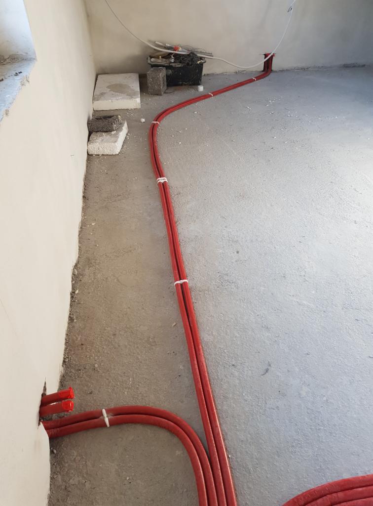 Czerwone rury instalacyjne, spięte białymi opaskami, prowadzone po surowym betonowym podłożu wzdłuż ściany w trakcie montażu.