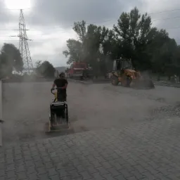 Pracownik z zagęszczarką płytową ubija podłoże z kruszywa pod przyszłą nawierzchnię, w tle koparko-ładowarka i ciężarówka, słup wysokiego napięcia.