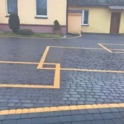 Ułożona, szara kostka brukowa z żółtymi obramowaniami kwadratów na podjeździe przed budynkiem.