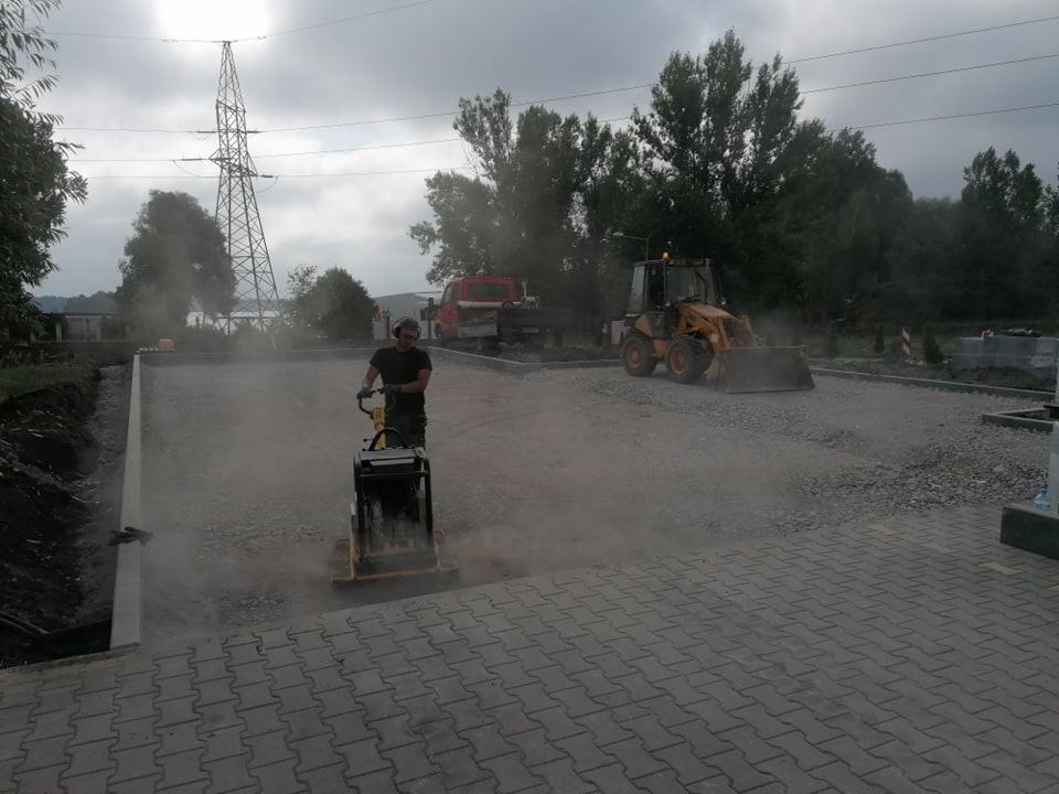 Pracownik z zagęszczarką płytową ubija podłoże z kruszywa pod przyszłą nawierzchnię, w tle koparko-ładowarka i ciężarówka, słup wysokiego napięcia.