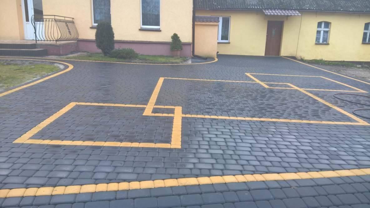 Ułożona, szara kostka brukowa z żółtymi obramowaniami kwadratów na podjeździe przed budynkiem.