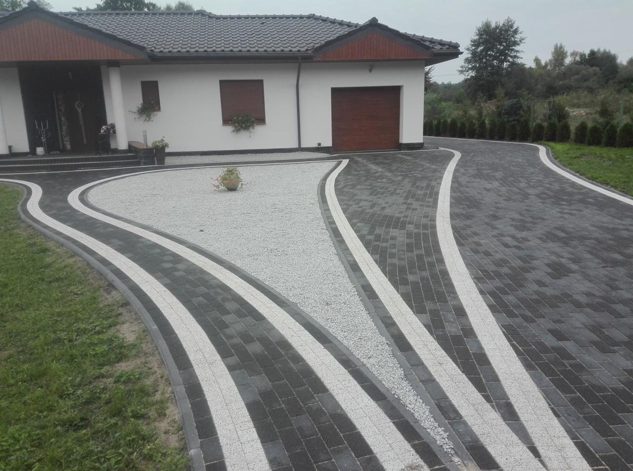 Podjazd do domu z kostki brukowej w odcieniach szarości, z białymi pasami i wypełnieniem żwirowym, widok z góry, dom w tle.