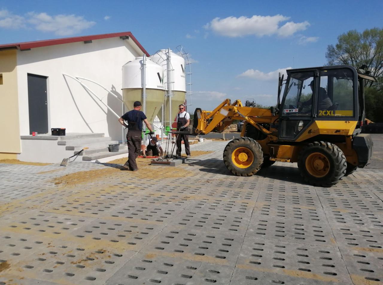 Pracownicy w Grubnie układają szarą ażurową kostkę brukową na podsypce piaskowej, widoczna żółta ładowarka i budynek gospodarczy z silosami w tle.