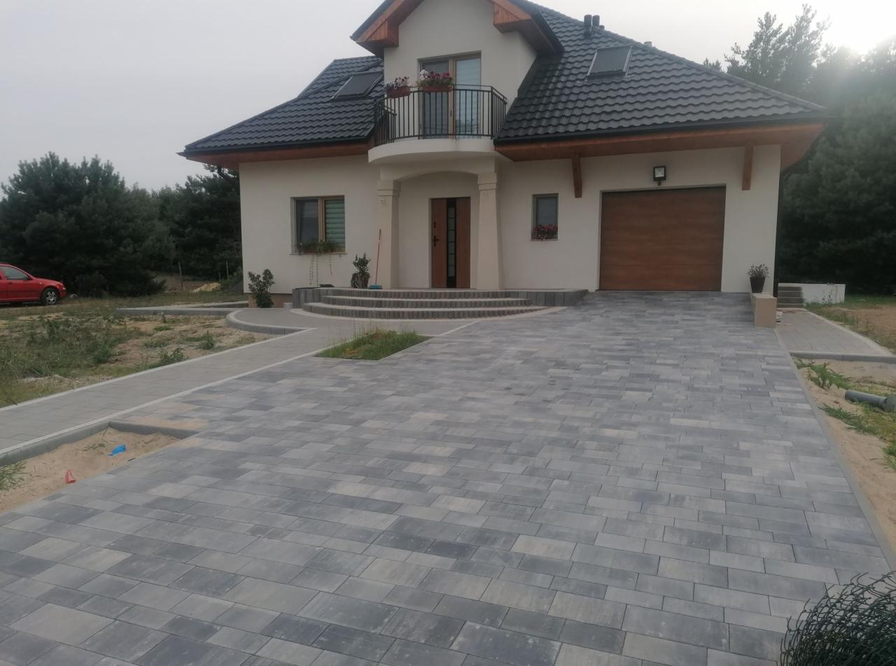 Nowoczesny dom jednorodzinny z podjazdem wyłożonym szarą kostką brukową w geometryczny wzór, z widocznymi schodami wejściowymi i fragmentem ogrodu.