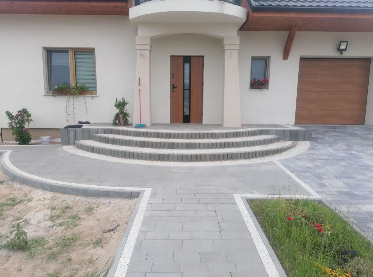 Nowoczesny podjazd z szarej kostki brukowej prowadzący do domu z drewnianymi drzwiami i schodami z zaokrąglonymi krawędziami, widoczne rabaty kwiatowe i trawnik.