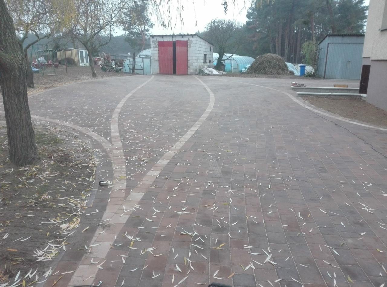 Szeroki podjazd z czerwonej kostki brukowej, z białymi pasami wyznaczającymi ścieżkę, zasypany opadłymi liśćmi. W tle widoczny budynek gospodarczy z czerwonymi drzwiami, drzewa i fragment domu.