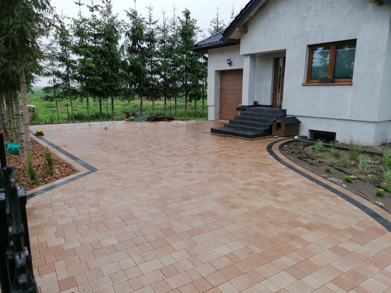 Beżowa kostka brukowa ułożona na podjeździe przed domem z szarym tynkiem, obrzeża z ciemnej kostki, w tle rząd świerków i zielone pole, widoczny fragment ogrodzenia.