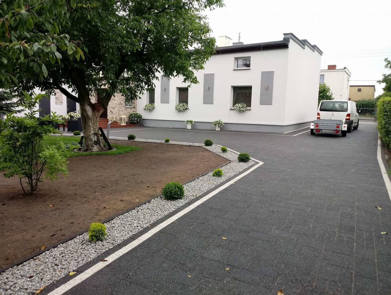Ukończony podjazd z ciemnej kostki brukowej z białymi liniami, obrzeża z szarego grysu, trawnik z przygotowaną ziemią, małe krzewy, biały budynek z kwiatami w oknach i zaparkowany biały samochód...