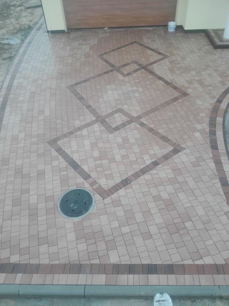 Ułożona kostka brukowa w odcieniach beżu i brązu z geometrycznym wzorem kwadratów, studzienka kanalizacyjna wkomponowana w nawierzchnię, widok z góry.