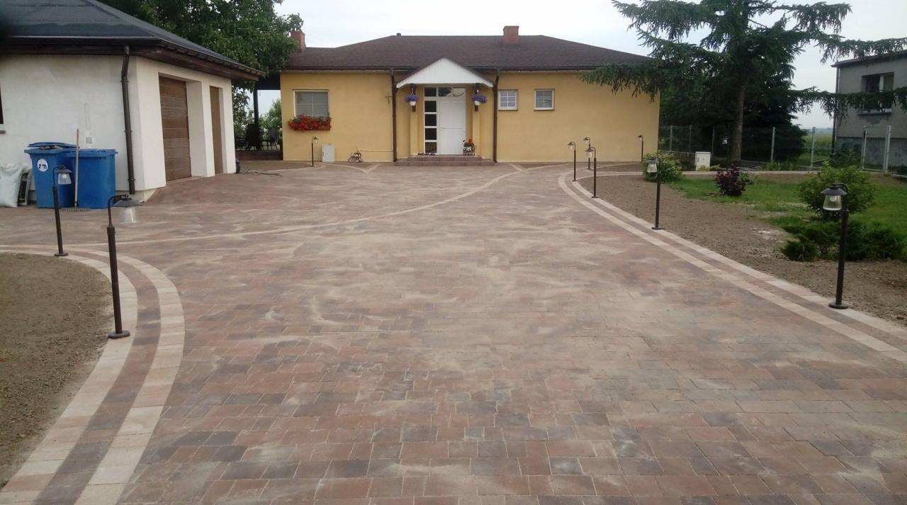 Nowo ułożona, wzorzysta kostka brukowa w odcieniach brązu i beżu tworzy rozległy podjazd do domu z żółtą elewacją, oświetlony przez niskie lampy ogrodowe wzdłuż krawędzi trawnika.