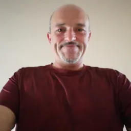 Portret uśmiechniętego mężczyzny z wąsami i brodą, ubrany w bordową koszulkę, na jasnym tle. Ujęcie z dołu.