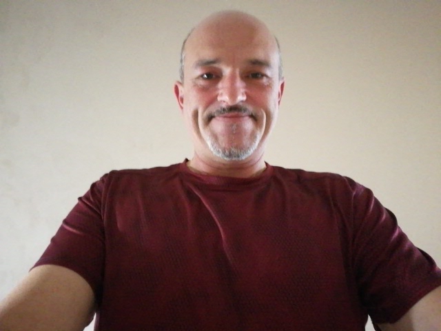 Portret uśmiechniętego mężczyzny z wąsami i brodą, ubrany w bordową koszulkę, na jasnym tle. Ujęcie z dołu.