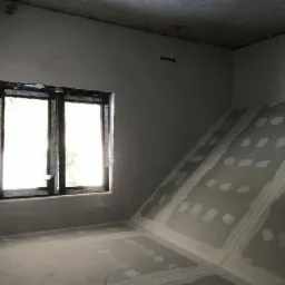 Surowe wnętrze poddasza ze skośną ścianą, przygotowane do dalszych prac wykończeniowych, widoczne łączenia płyt kartonowo-gipsowych, okno w czarnej ramie z folią ochronną.