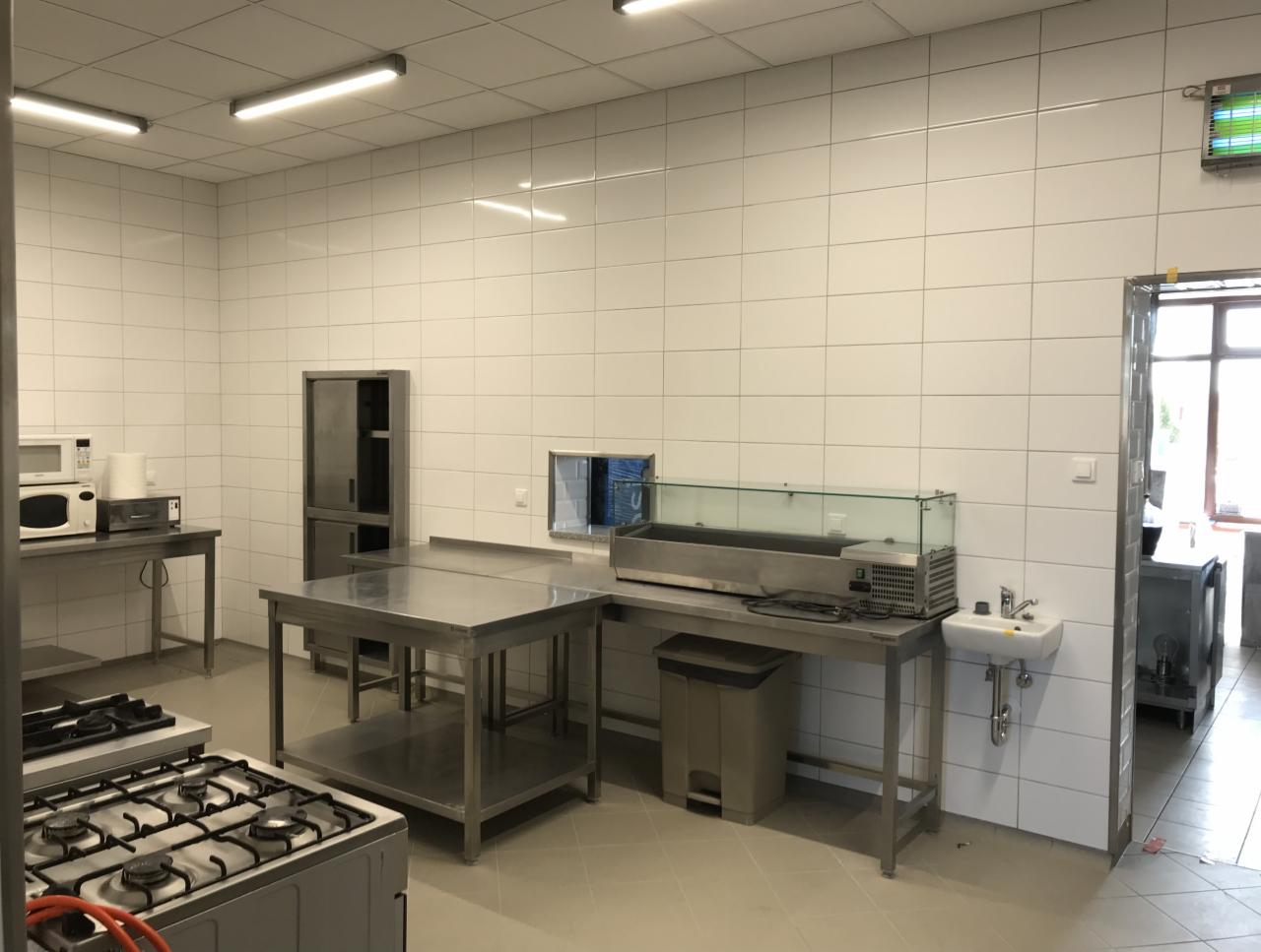 Wnętrze kuchni przemysłowej z białymi płytkami na ścianach, stalowymi stołami roboczymi, kuchenką gazową, szafką i małą umywalką.