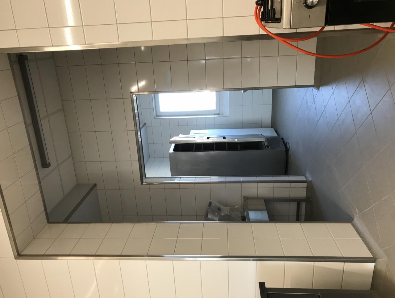 Wykończona kuchnia przemysłowa z białymi kafelkami na ścianach i szarymi płytkami na podłodze, widoczna metalowa szafka chłodnicza pod oknem i fragment instalacji elektrycznej.