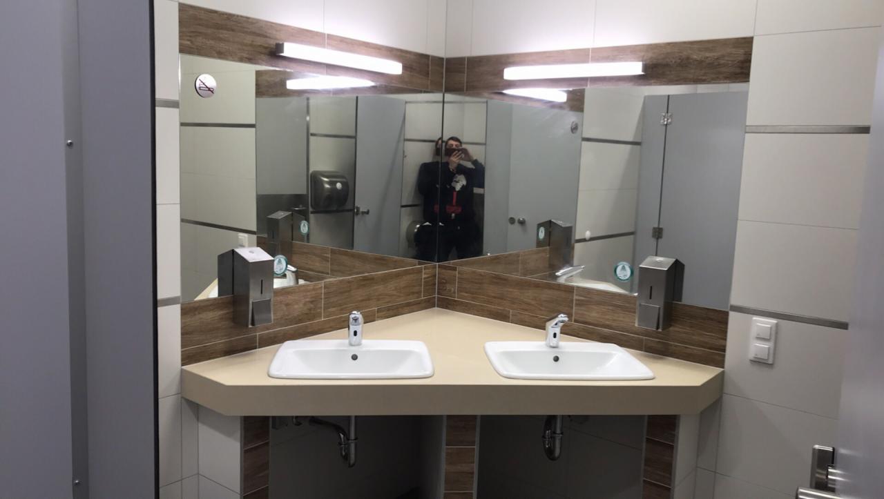 Publiczna toaleta z dwoma umywalkami, dużym lustrem i dozownikami mydła w metalowych obudowach. W lustrze odbija się osoba robiąca zdjęcie.