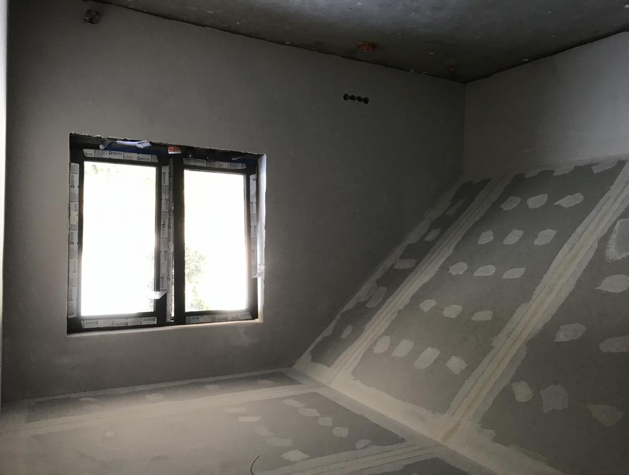 Surowe wnętrze poddasza ze skośną ścianą, przygotowane do dalszych prac wykończeniowych, widoczne łączenia płyt kartonowo-gipsowych, okno w czarnej ramie z folią ochronną.