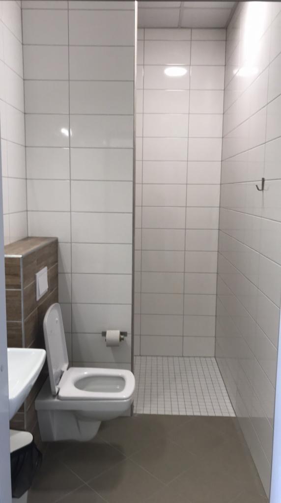 Nowoczesna toaleta z białymi płytkami, prysznicem bez brodzika i szarą podłogą. Widoczny wiszący sedes z otwartą klapą, umywalka i uchwyt na papier toaletowy.