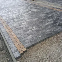 Ułożona z szarej kostki brukowej ścieżka z obramowaniem z jasnej kostki, prowadząca do kamiennych filarów. Widoczne niedokończone fragmenty i podsypka z kruszywa.